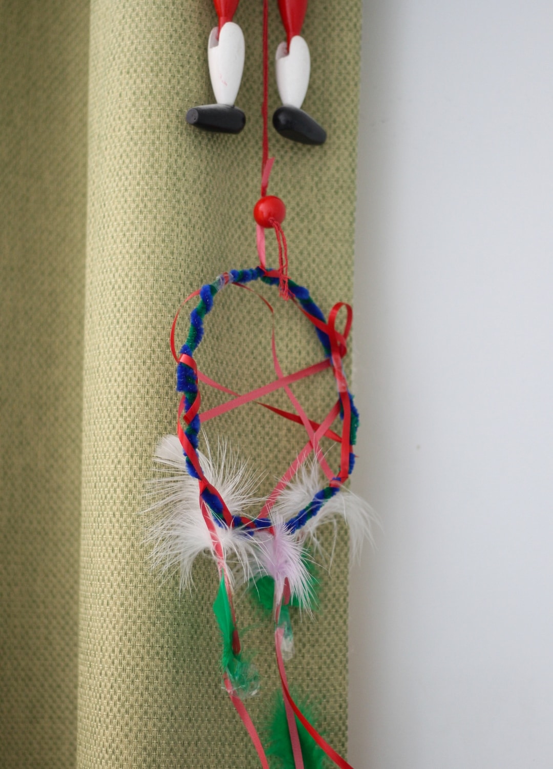 A dream catcher made by a child, hanging down from a curtain with the legs of a wooden toy above