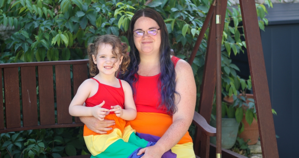 A woman and small girl dressed in matching rainbow dresses, sitting on a bench swing. Rainbow colours for support of adult autism diagnosis.