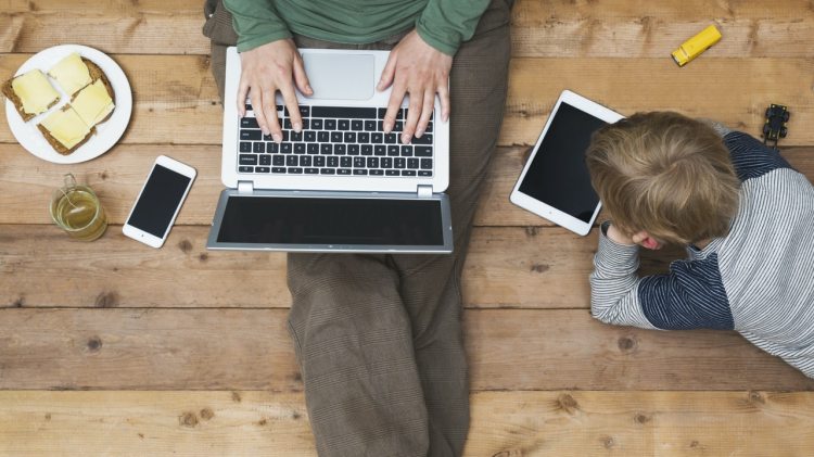 Image of person with laptp on lap. Child playing on tablet next to them.